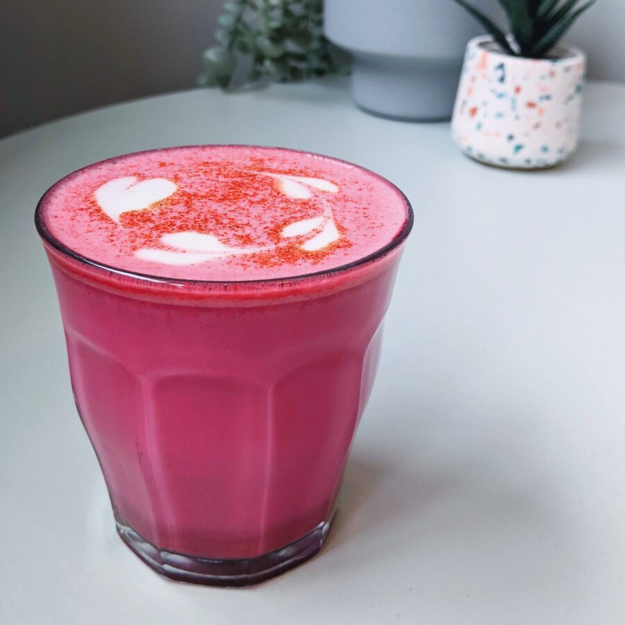 A pink beetroot latte in a glass on a pale table, with plants behind.